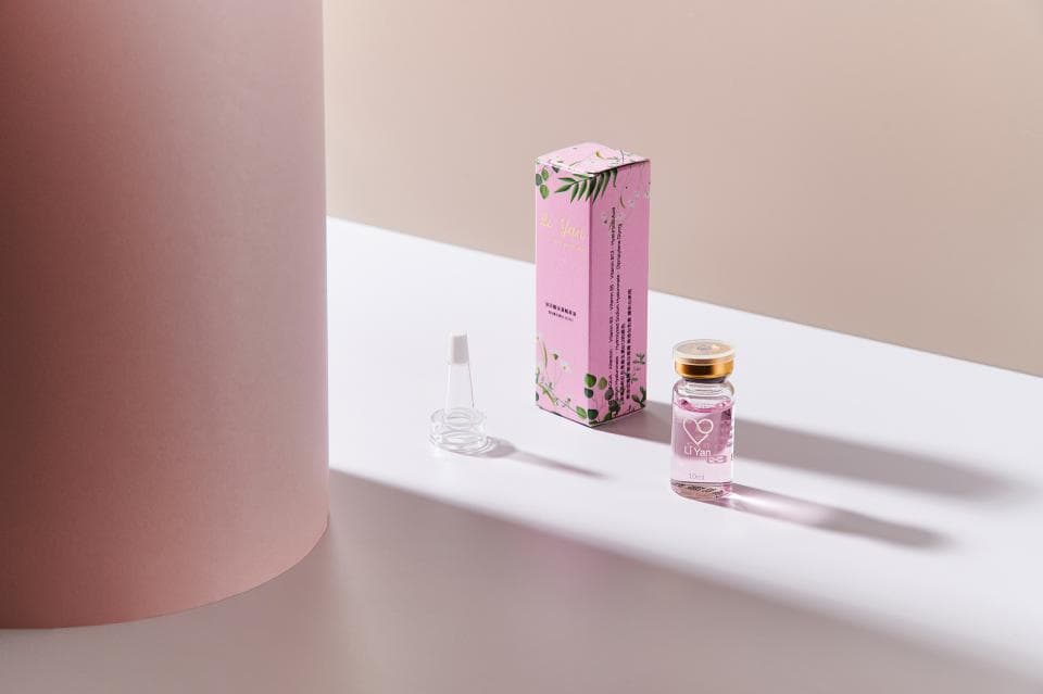 這張照片是一瓶粉色玻璃瓶裝的美容液,瓶子上面印著愛心的圖案,旁邊是一盒粉紅色的包裝盒,盒子上印著花朵和葉子的圖案,看起來很可愛。