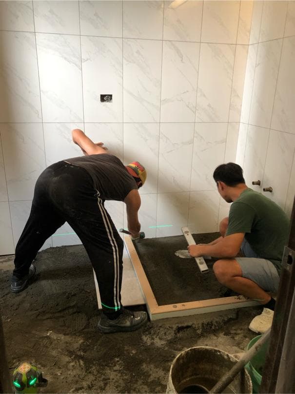 這張照片是一個正在施工的浴室,最重要的部分是地面上的水泥和木板,一個工人正在使用水平儀來確保水泥板的平整。
