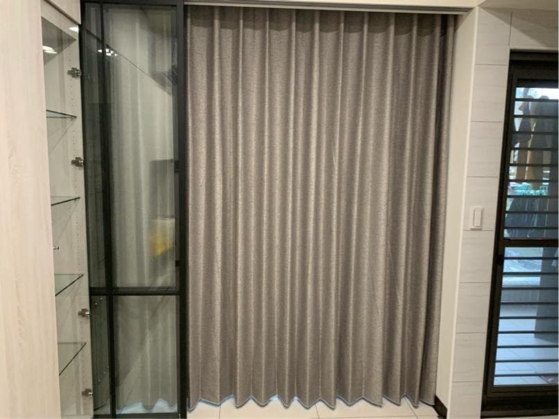 1) 照片顯示一個客廳的角落,有一個玻璃櫃和一扇灰色窗簾,旁邊的牆上有個白色開關,窗戶可以看到外面的景色