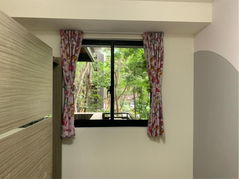 1) 照片顯示一個房間的一角,裡面有一扇窗戶,窗戶上掛著粉紅色的窗簾,窗外可以看到樹木,房間的牆壁是淺灰色的,房間裡還有一部分木質的牆面