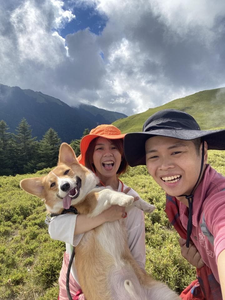 ## 照片資訊:
**1) 照片描述:** 照片中,兩位年輕女性在山區戶外背景中與一隻可愛的柯基犬互動,其中一位女生抱著柯基犬,而另一位女生則站在旁邊,三人皆露出開心的笑容