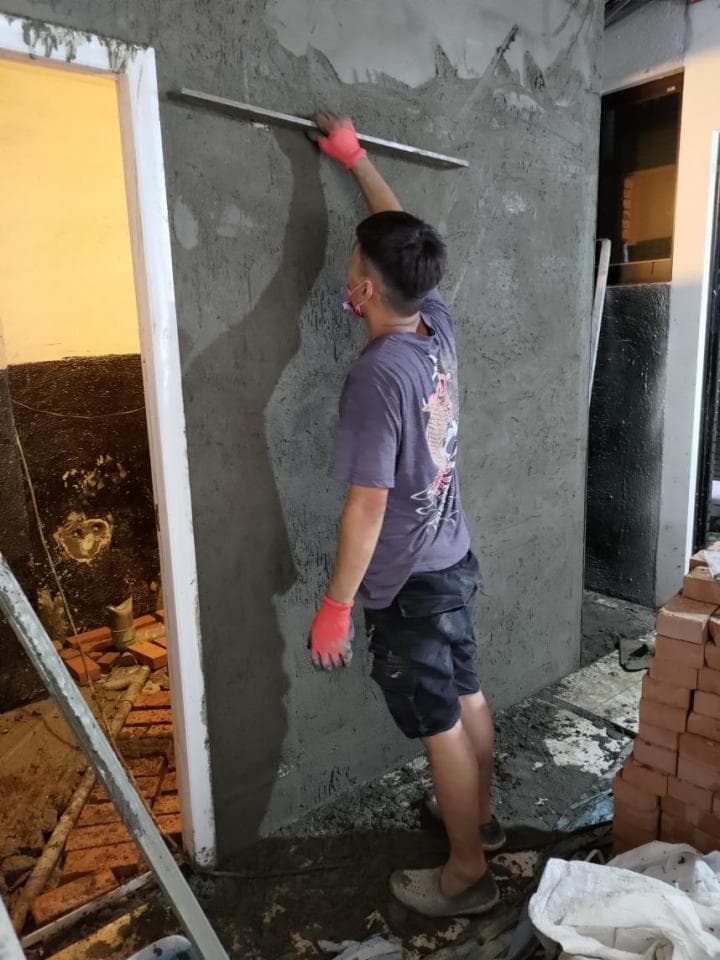 1) 照片顯示一位身穿灰黑色T恤和黑色短褲的男性工人,正在使用抹刀抹水泥,牆上已塗抹了一層水泥,他站在正在施工的房間裡,房間的地板尚未完成,地上散落著一些磚塊