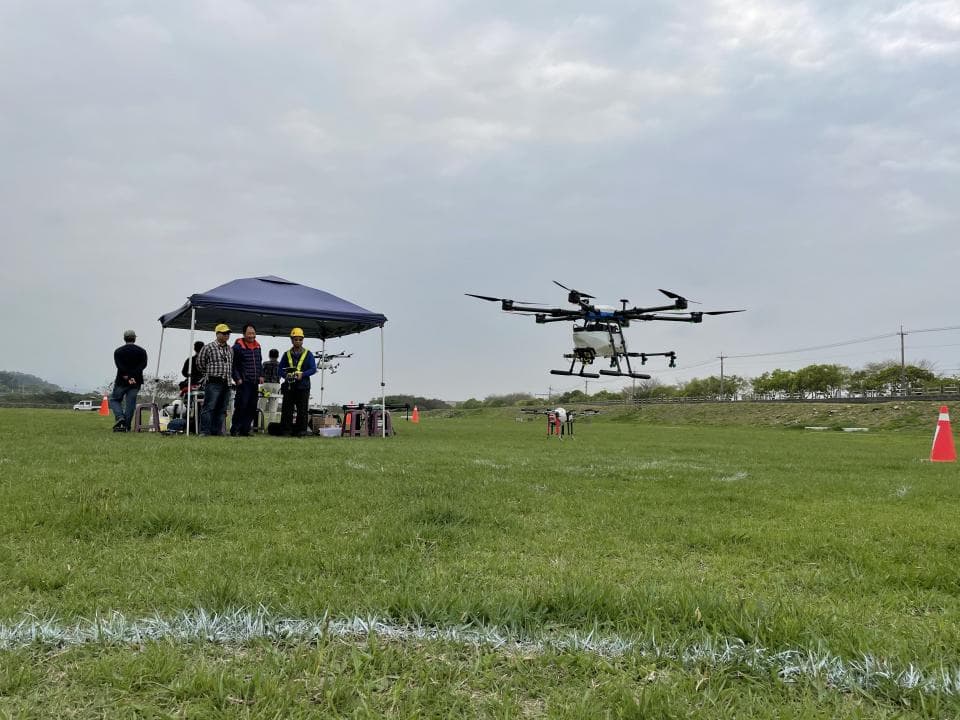 這張照片是一架無人機正在空中飛行,它看起來很像一架小型直升機。它有四個旋翼,以及一個掛在底部的白色容器。它正飛過一片草地,地上有一群人正在觀看。