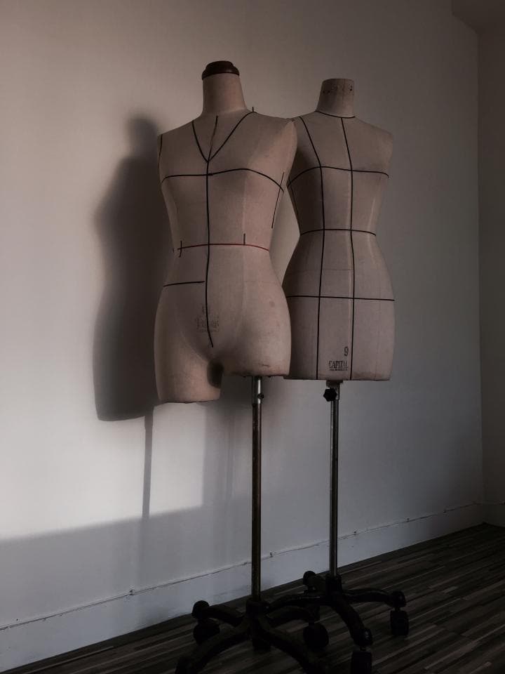 這張「兩個裁縫人臺」的照片是,兩個裁縫人臺,一個是女性的,一個是男性的,都立在白色牆壁前,人臺上都有標記線,它們都放在金屬支架上,並配有輪子。