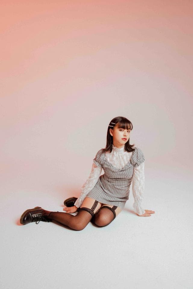 1) 照片中是一位年輕女子,穿著格紋洋裝,坐在白色背景前,她的腿上穿著黑絲襪,並搭配黑色短靴