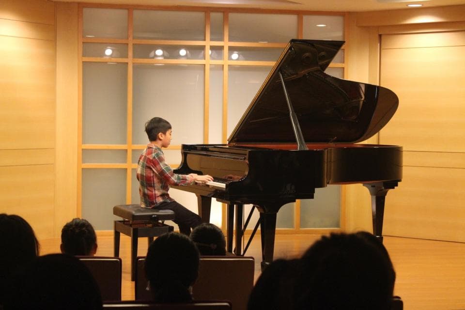 這張照片是一個年輕人正在彈鋼琴。他坐在鋼琴前,雙手放在鍵盤上,專注地演奏著。鋼琴是黑色的,非常大,看起來很昂貴。房間的背景是木板牆,給人一種溫馨舒適的感覺。