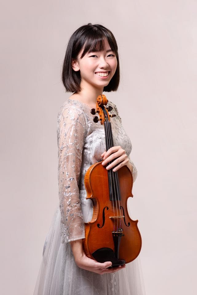 1) 照片中是一位穿著白色蕾絲洋裝的年輕女性,她正拿著一把小提琴,站在淡色的背景前,並露出微笑