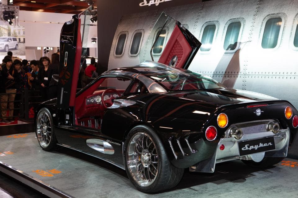 這張「Spyker C8」的照片是一輛黑色的跑車,它有兩個鷗翼式車門和一個紅色內飾,背景是一架飛機。