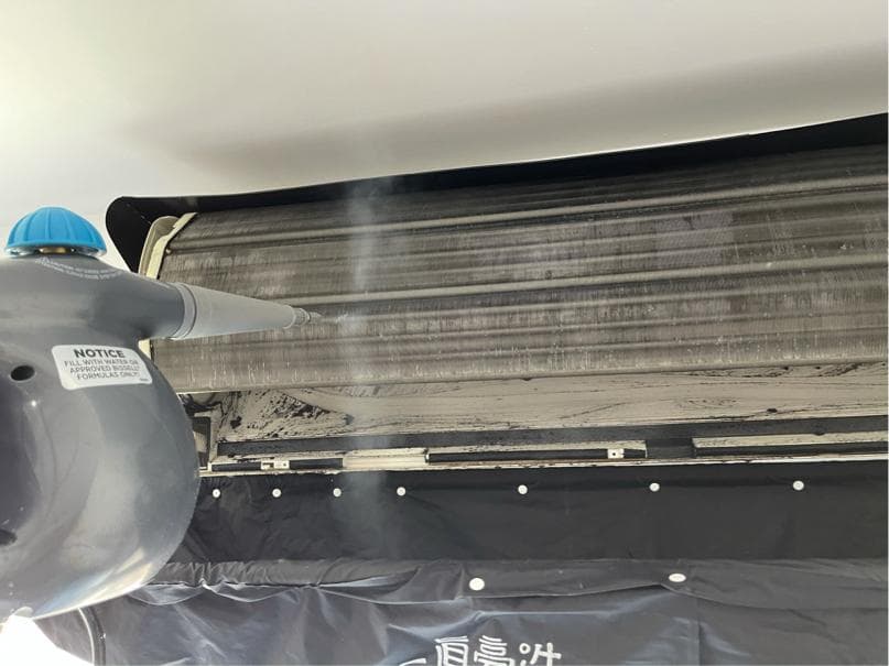 1) 照片顯示一個正在使用蒸汽清洗空調冷凝器的過程,蒸汽機正噴出蒸汽,而冷凝器下方則覆蓋著黑色的布