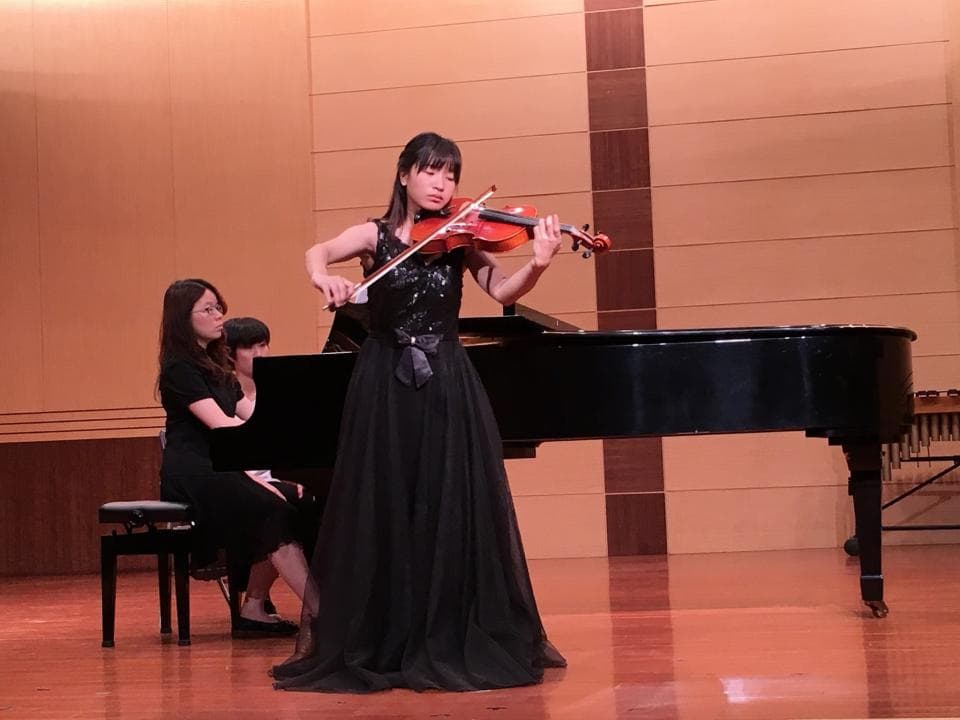 這張照片是一位演奏小提琴的年輕女性。她穿着黑色長裙,正在舞台上演奏小提琴。她身後放着鋼琴,看起來像是音樂會的演出。
