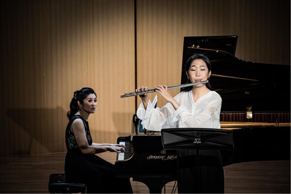 這張「兩位女性音樂家在舞臺上表演」的照片是:一位女士坐在鋼琴前彈奏,另一位女士站在鋼琴旁吹奏長笛。她們都穿著正式服裝,展現出專業的音樂家風範。