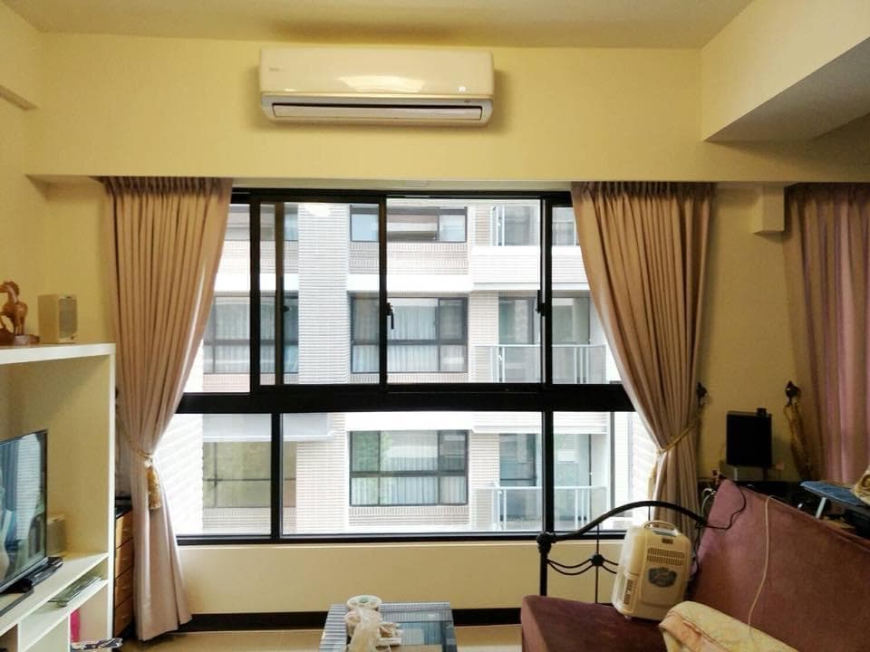 1) 照片顯示一間客廳,房間的牆壁是米白色的,牆上裝了冷氣,窗戶採落地窗設計,窗戶旁裝了窗簾,客廳擺放了一張沙發,沙發上有抱枕和毯子