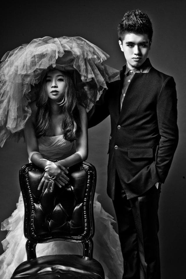 這張「婚紗照」的照片是一對情侶,穿著西裝和婚紗,男方站在女方身旁,女方坐在黑色皮椅上,背景是灰色的牆壁。