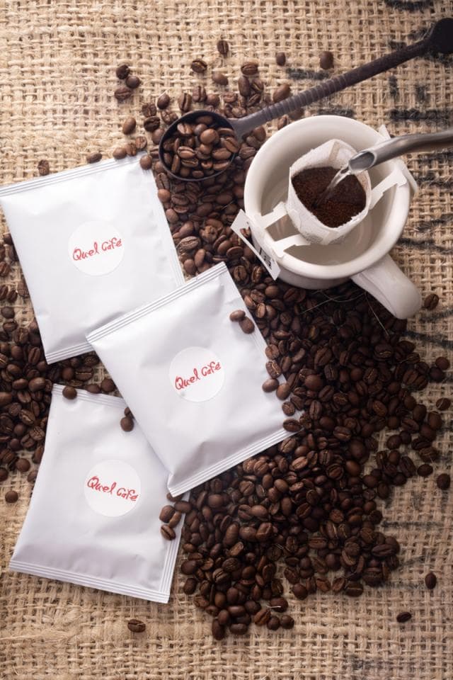 這張照片是一包咖啡豆,豆子散落在粗麻布上,旁邊有三個咖啡包和一個咖啡壺。