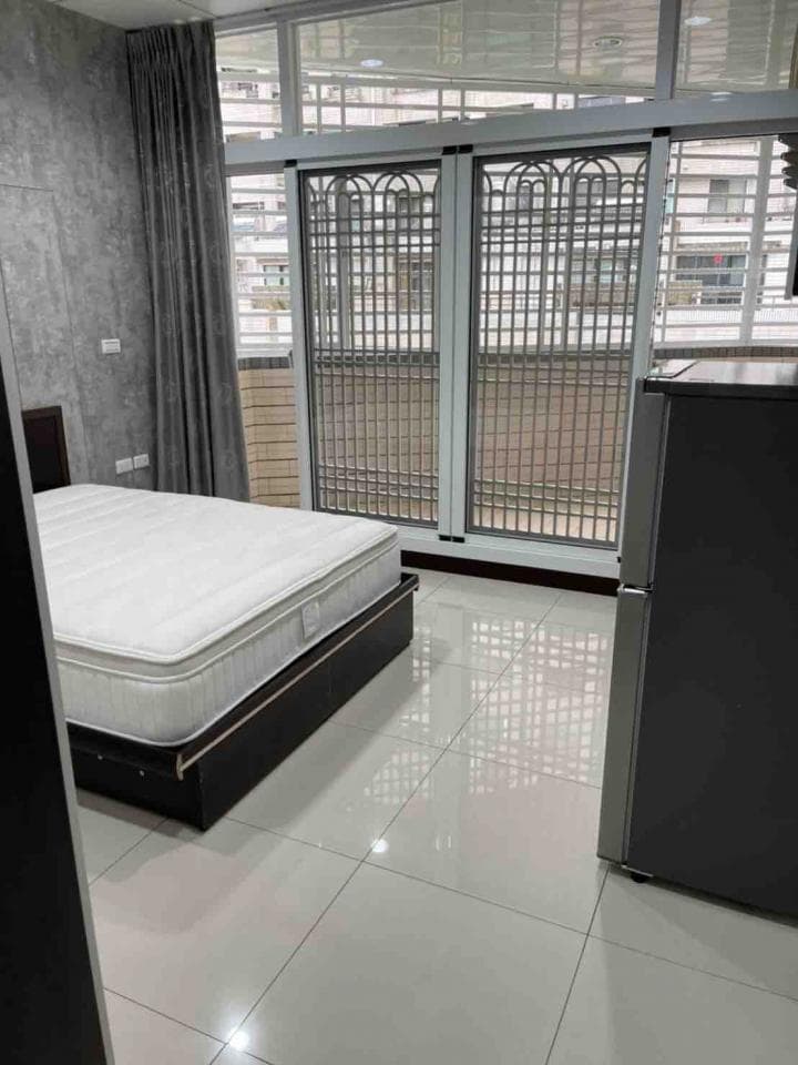 1) 照片顯示一個白色床墊放在一個黑色的床架上,床架旁邊是一個黑色的冰箱,床的後方有一個落地窗,落地窗外面是陽台,落地窗上是百葉窗