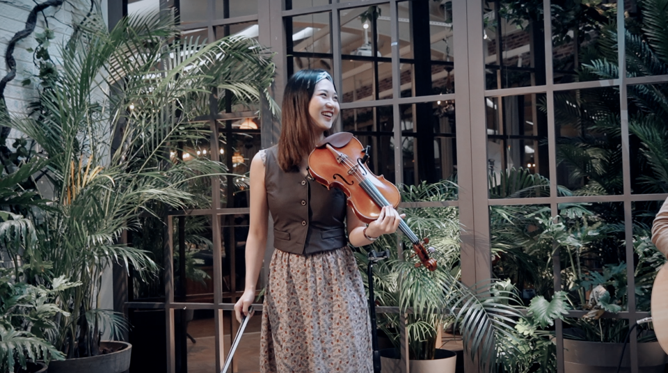 這張照片是一個年輕女子在演奏小提琴。她穿著一件深色的背心和一條印花裙,看起來很開心。她右手拿著小提琴,左手在琴弓上。背景是綠色的植物和玻璃門。