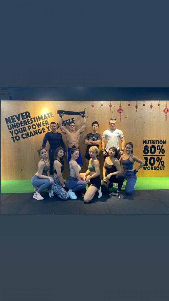 這張健身房照片是一群朋友在健身房裡,他們一起完成了訓練,並開心地在健身房前拍照留念。他們身穿運動服,展現出健康自信的活力,牆上寫著「營養佔80%,運動佔20%」,鼓勵大家重視健康生活。