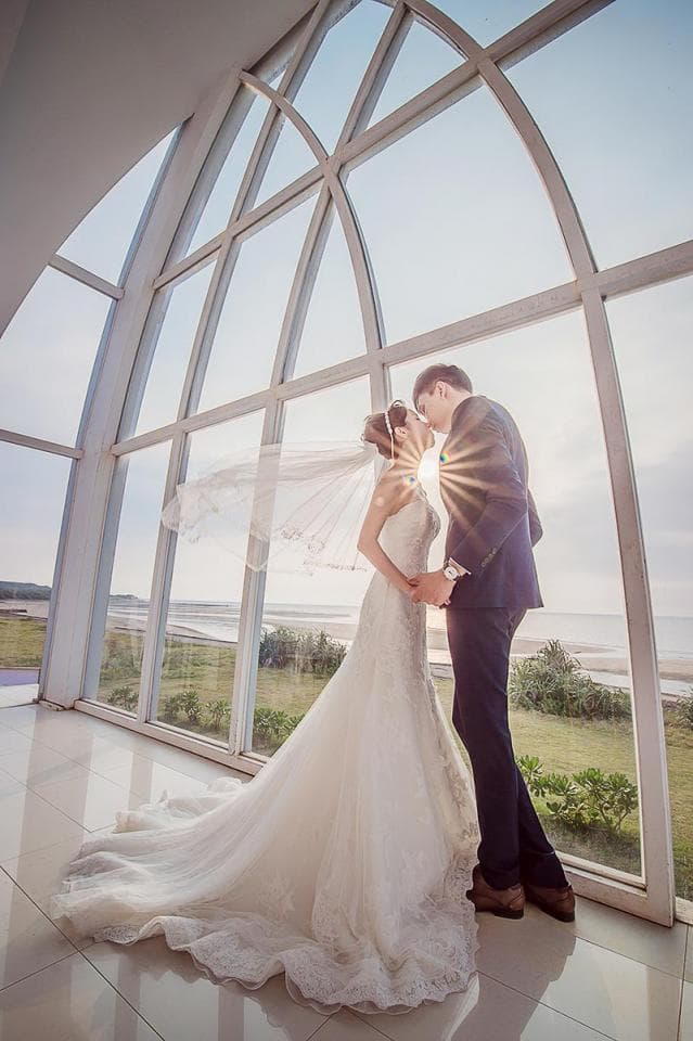這張婚禮的照片是一對新人站在巨大的落地窗前,新娘穿著白色婚紗,新郎穿著深色西裝,他們深情相擁,背景是蔚藍的天空和美麗的風景。