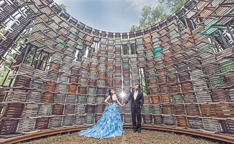 這張照片是一對新人站在由許多木板組成的圓形建築物中,新郎穿著西裝,新娘穿著藍色禮服,他們手牽著手,看起來非常幸福。