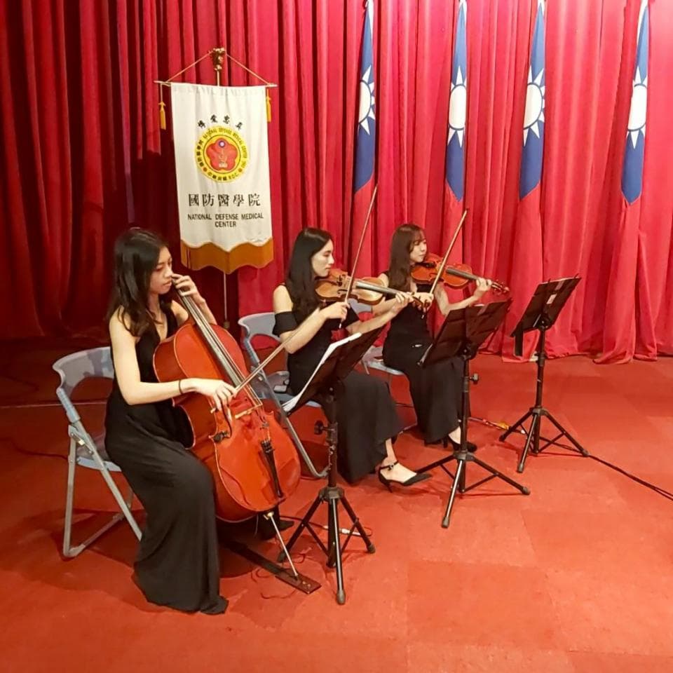 這張「三位女性演奏家在紅色布幕前演奏」的照片是,她們身穿黑色禮服,分別演奏大提琴、小提琴,並在演奏臺上使用譜架。