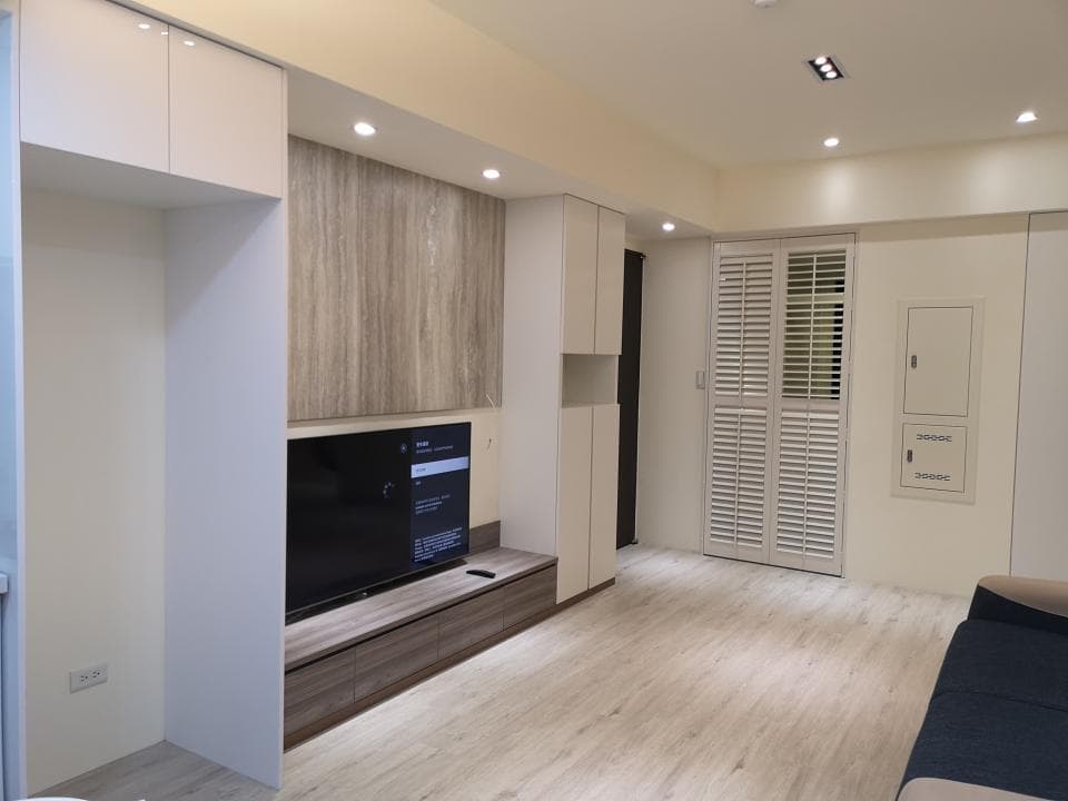 這張照片是一個現代化的客廳,最重要的是電視牆,它由木紋裝飾面板、電視機和下方收納櫃組成。牆面採用淺色調設計,讓空間顯得寬敞明亮。