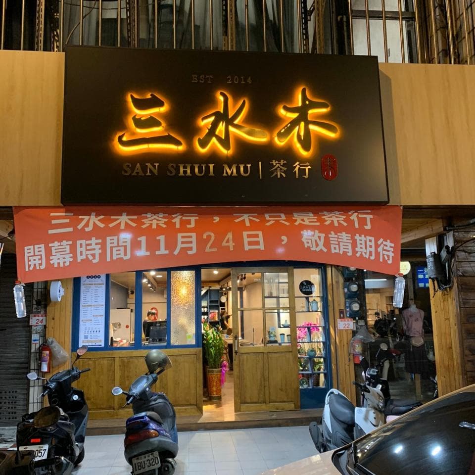 這張「三水木茶行」的照片是一家茶行,招牌上寫著「三水木茶行」,門前有兩臺機車,還有紅色布條寫著「三水木茶行,不只是茶行,開幕時間11月24日,敬請期待」。