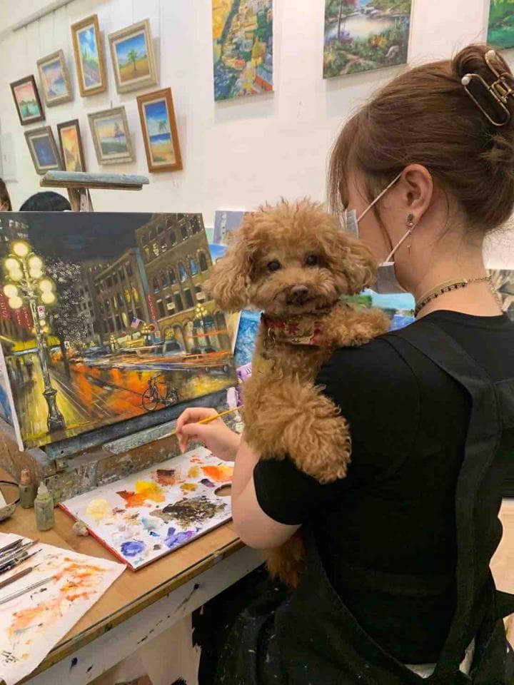 這張照片是一個女生正在畫畫,她旁邊還有一隻可愛的狗狗陪著她,牠的毛色是淺棕色,看起來很溫順。