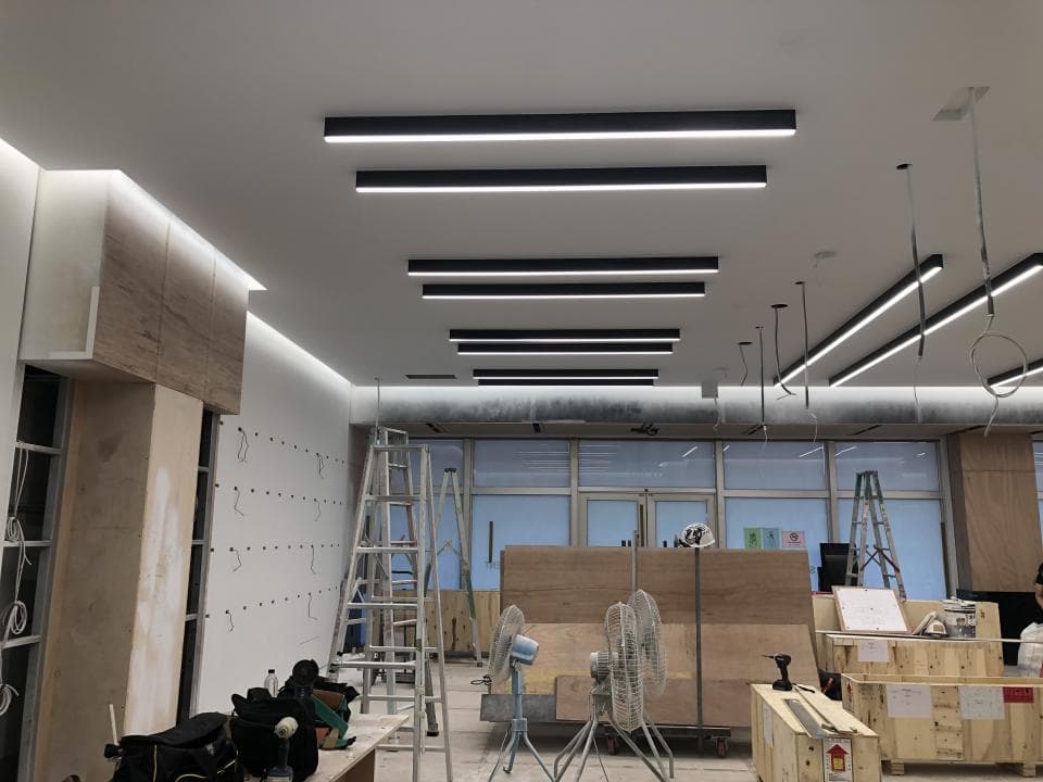 1. 照片顯示一個室內空間,天花板裝有黑色線性燈,空間內有木板,梯子,風扇,部分空間牆壁正在粉刷