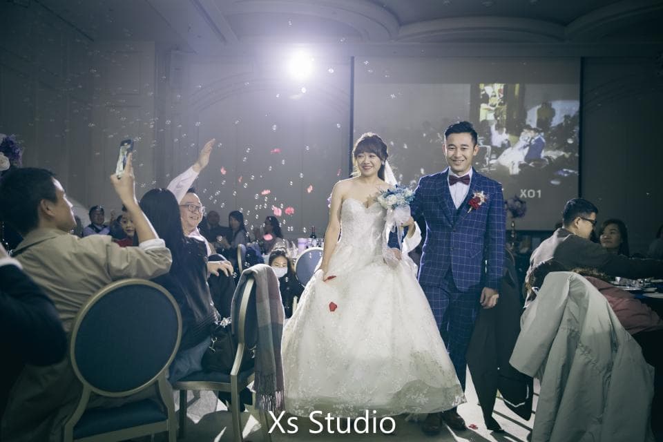 這張婚禮的照片是一對新人走在紅毯上,新郎穿著藍色格紋西裝,新娘穿著白色婚紗,他們在眾人的祝福下步入禮堂。