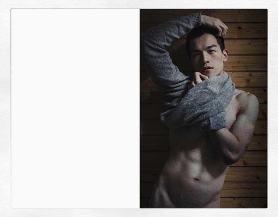 這張照片是一個年輕男子半裸著上半身,他用手抓著一件灰色毛衣,露出精壯的肌肉和堅毅的眼神。