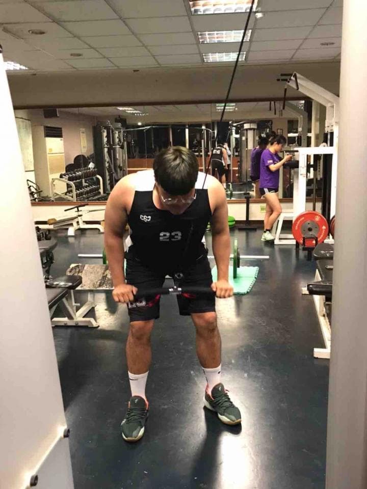 這張照片是一個年輕男子在健身房使用拉力器鍛鍊背部肌肉。 他穿著黑色運動服和綠色的運動鞋,正專注地拉動拉力器。 他周圍有一些健身器材,但他的背部肌肉才是照片的重點。