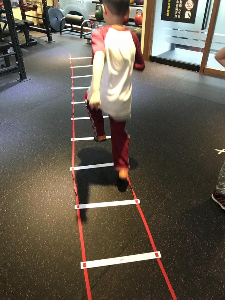 這張照片是一位年輕女子,她穿著白色襯衫和棕色寬腿褲,站在木頭椅子上,背景是白色牆壁和綠色植物。