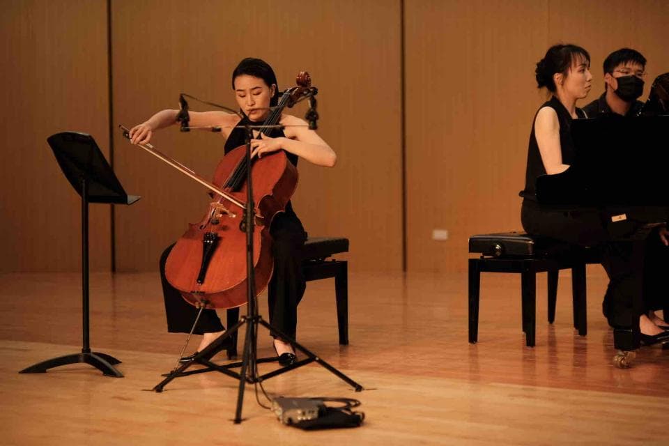 這張「音樂表演」的照片是一名女演奏家在演奏大提琴,她身穿黑色連身裙,正專注地拉著琴。她的右手輕輕撫摸琴絃,左手則按住琴頸,展現出精湛的琴藝。在她身後,兩位觀眾坐在椅子上觀賞表演。