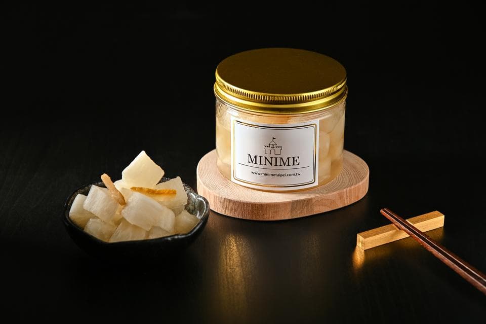 這張「醃製蘿蔔」的照片是一個玻璃罐裝的醃製蘿蔔,上面貼著「MINIME」的品牌標籤,旁邊放著一碗醃製蘿蔔和兩根筷子,看起來很美味。