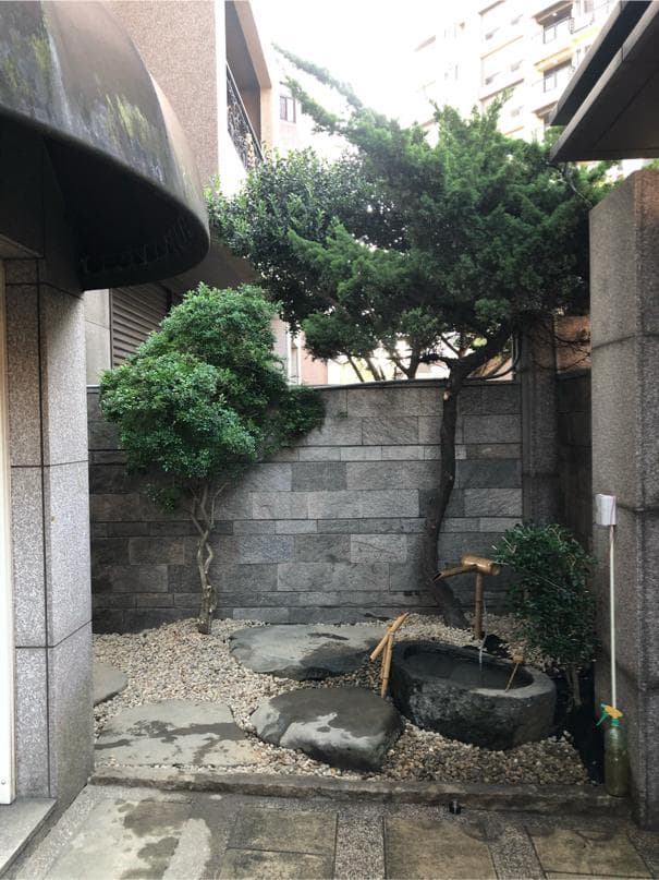 這張照片是一處小巧的庭院,其中最引人注目的是一個由石頭砌成的方形水池,水池中有一個木質水管,水管上有一個木質水龍頭,水龍頭正滴著水,水池周圍佈滿了鵝卵石。