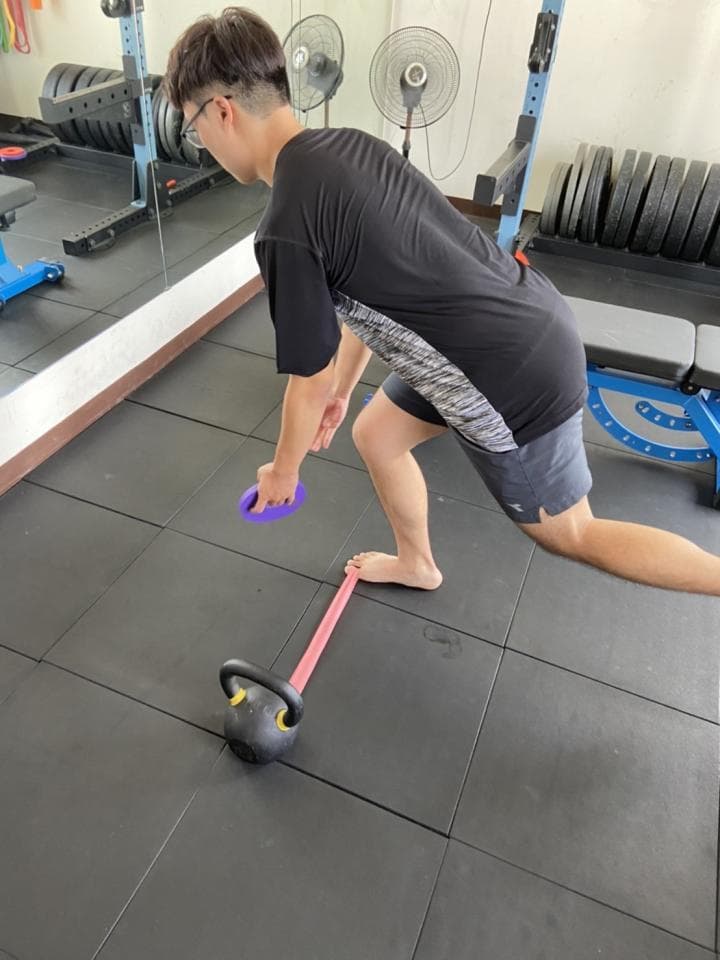 這張照片是一個男人在健身房裡做運動。他正在做一個單腳舉重訓練,右腳踩在粉紅色的阻力帶上,左手拿著紫色的滑輪,看起來很專注。