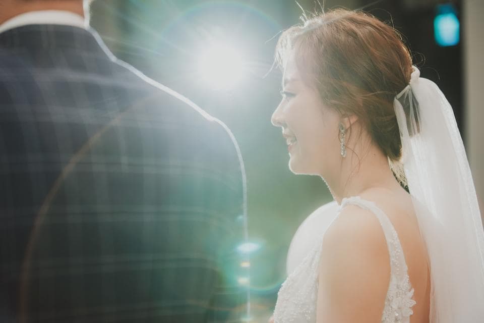 這張婚禮照片是一個新娘,她穿著白色婚紗,頭戴白紗,正在微笑,她的丈夫站在她背後。