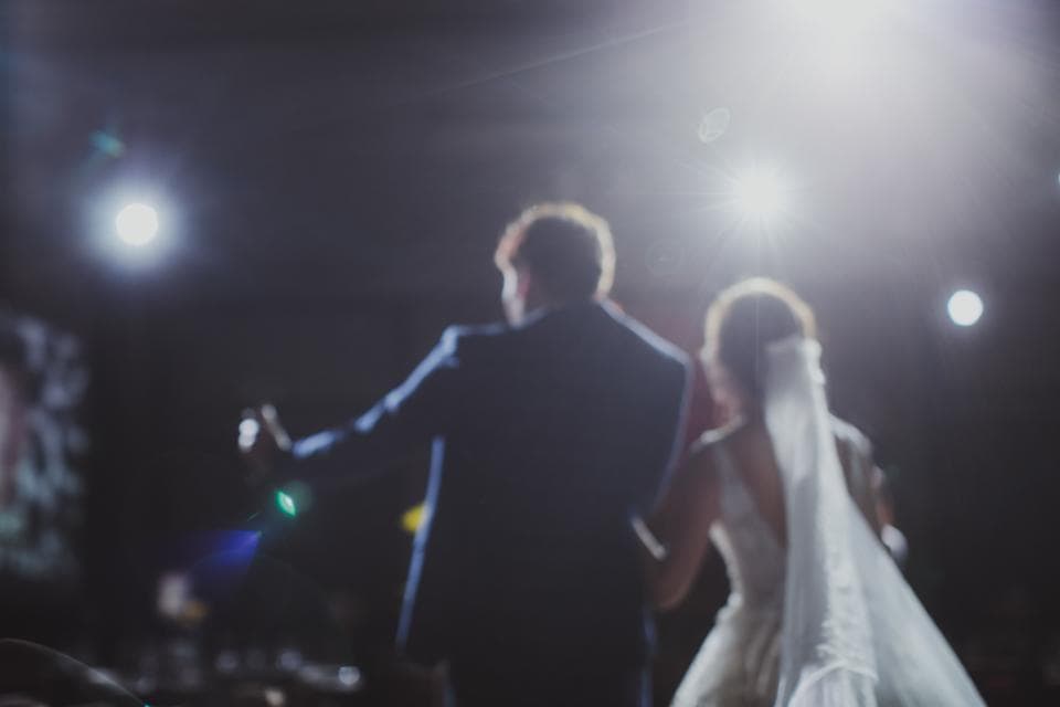 這張照片是一對新人站在臺上,他們背對著鏡頭,看不清他們的臉,但可以看見他們穿著婚禮服裝,男方穿著西裝,女方穿著婚紗。他們在聚光燈下,看起來非常幸福。