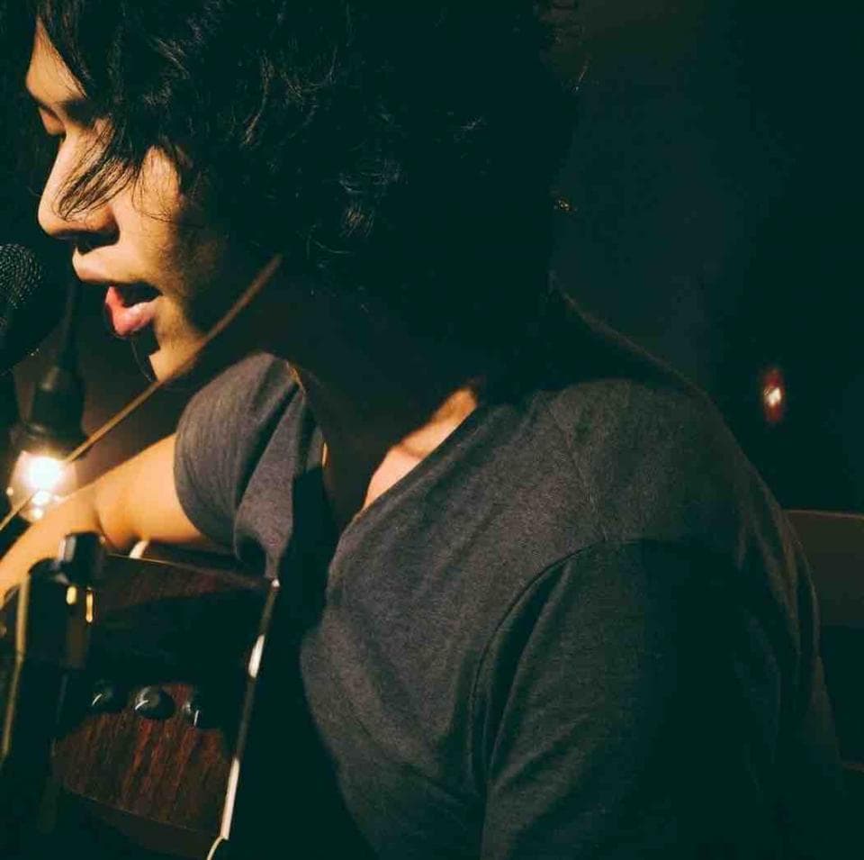 這張照片是一個男人正在唱歌,他穿著灰色的 T 恤,頭上戴著耳機,背景是一把吉他。