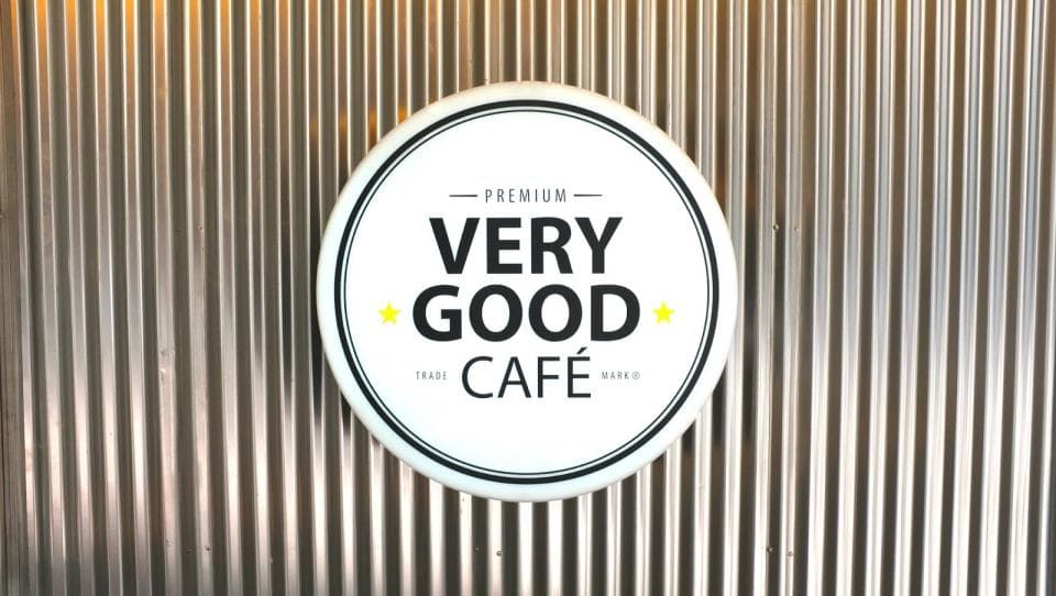 這張「咖啡店招牌」的照片是一個圓形的白色招牌,上面寫著「VERY GOOD CAFÉ」。招牌是貼在一個金屬牆上,牆上有很多垂直的線條。