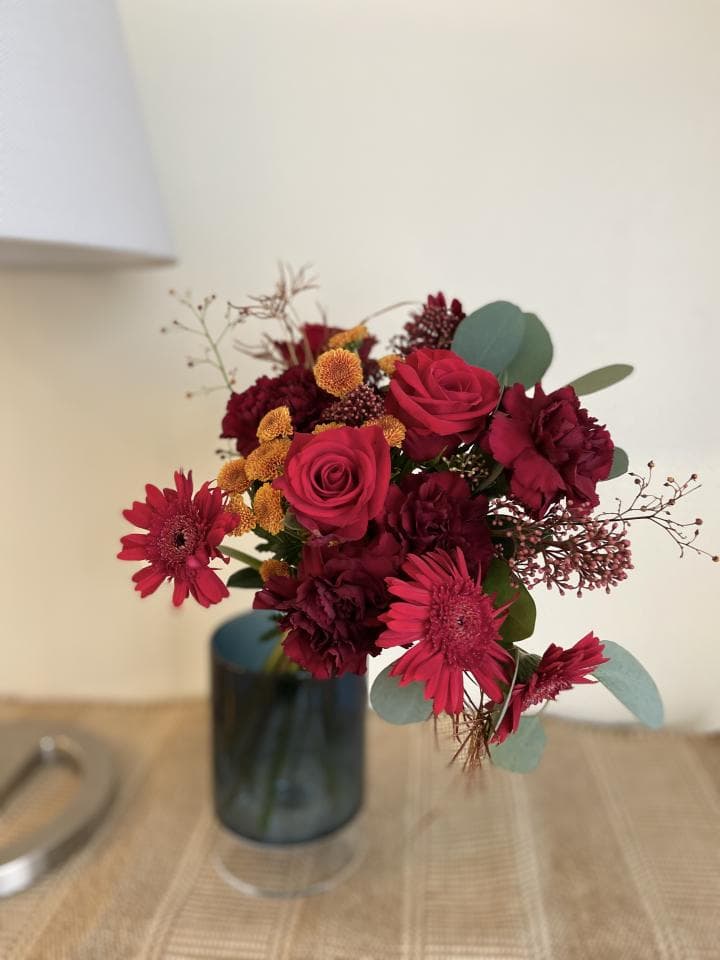 1) 照片中是一束紅色的花,花束裡面有玫瑰、菊花和一些其他綠色葉子,花束放在一個藍色的花瓶裡面,花瓶放在一個棕色編織的桌子上