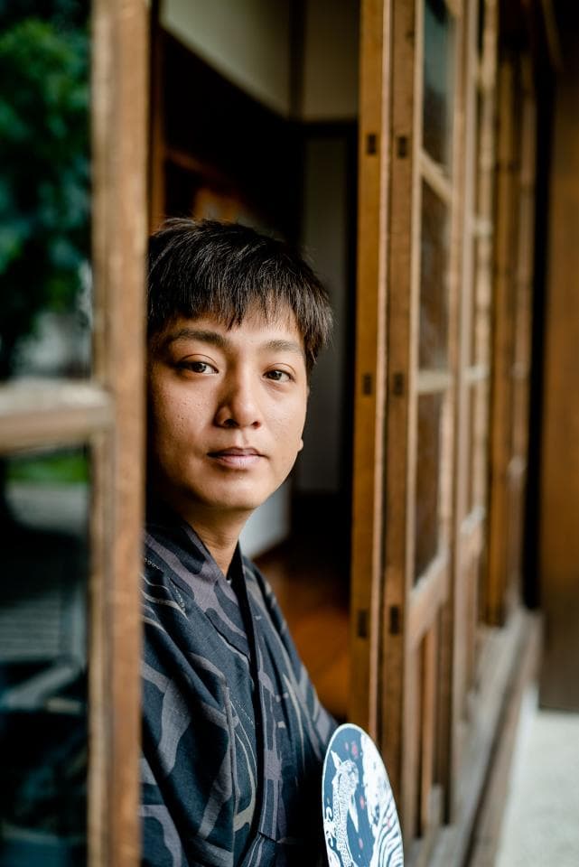 這張照片是一個年輕男子站在日式建築的窗戶旁,他穿著傳統的和服,看起來很沉靜。