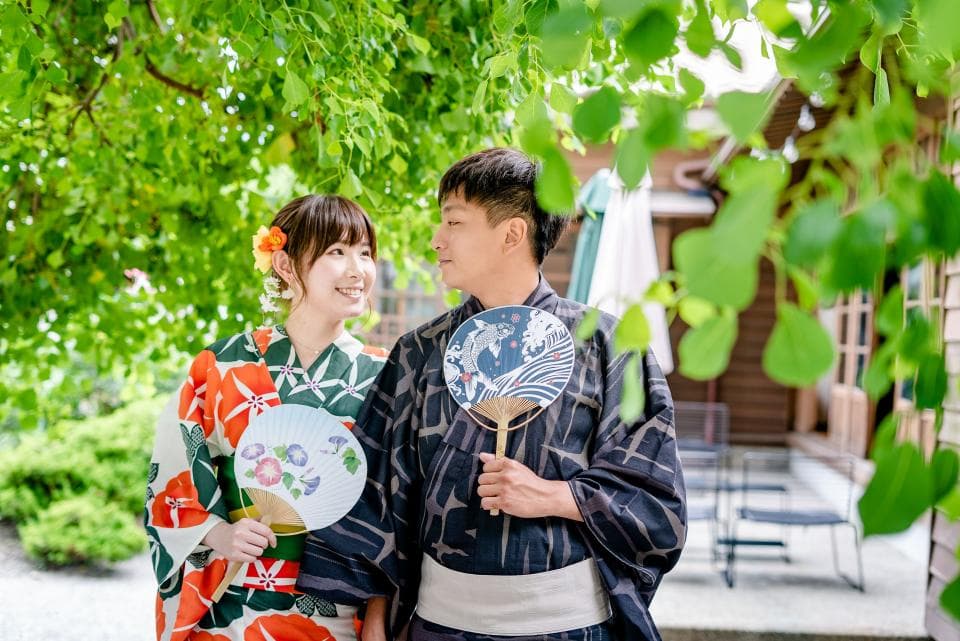 這張「穿著和服的男女」的照片是,一對男女在樹蔭下,穿著傳統和服,手持扇子,面帶微笑,背景是一間日式建築。