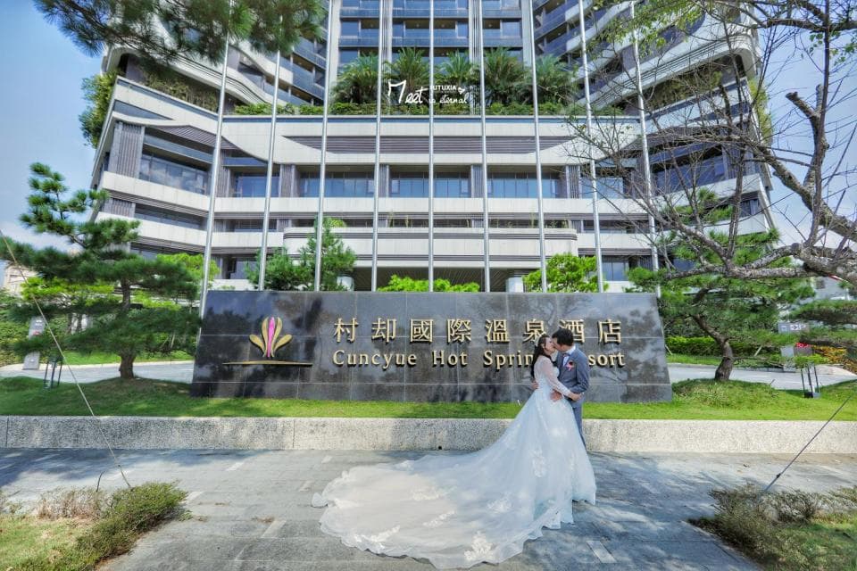 這張照片是 一對新人站在「村卻國際溫泉酒店」的招牌前,新娘穿著潔白的婚紗,新郎則穿著西裝,他們正幸福地擁吻。