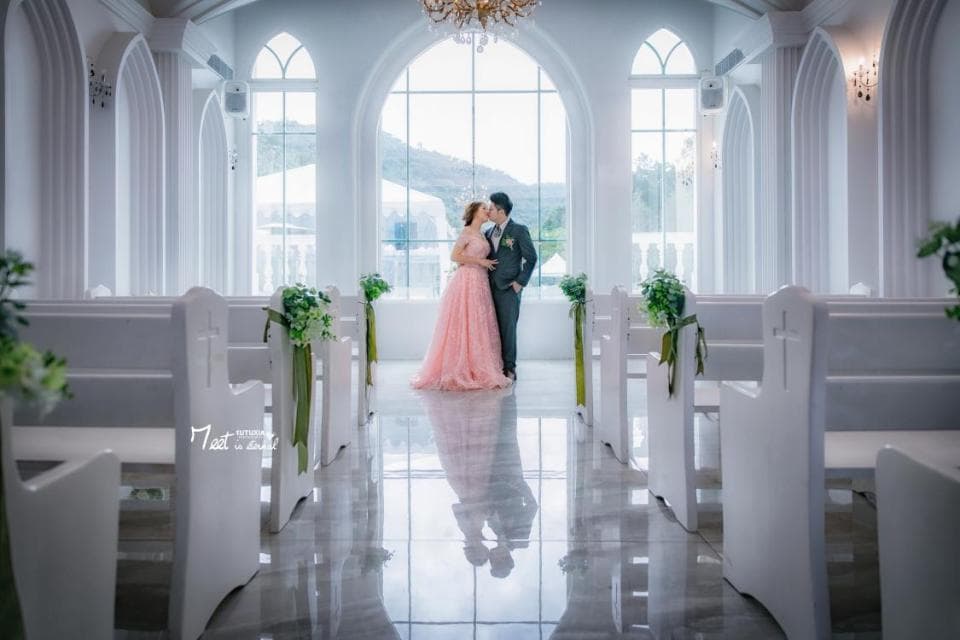 這張照片是一對新人正在教堂裡親吻,背景是白色教堂和綠色植物。他們身穿婚紗和西裝,看起來非常幸福。