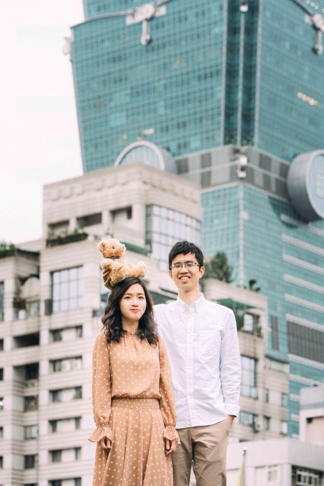 這張照片是一對情侶在城市中合照,女生頭上戴著一隻毛絨玩具熊,男生站在女生旁邊,他們看起來很幸福。背景是一棟高樓大廈,顯得十分熱鬧。