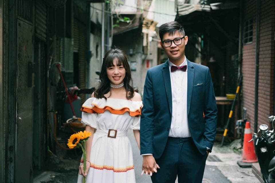 這張「婚紗照」的照片是一對情侶在巷子裡拍的。女孩穿著白色洋裝,男孩則穿著西裝,他們都很開心,手牽著手。