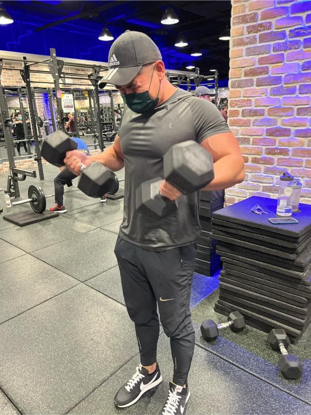 這張照片是一名男子在健身房舉啞鈴,他穿著運動服,戴著黑色帽子,表情專注。