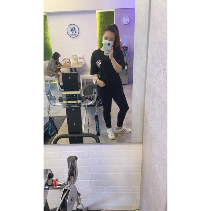 這張「健身房」的照片是一名女性站在健身器材前,鏡子裡映照出她的全身。她穿著黑色上衣和褲子,戴著口罩,正在使用健身器材。