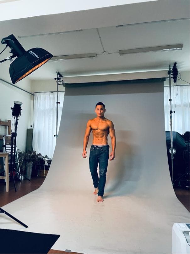 這張「攝影棚」的照片是,一個男性模特兒站在白色背景布前,準備拍攝。他穿著牛仔褲,露出上半身肌肉。照片中也包含攝影燈光和棚架。